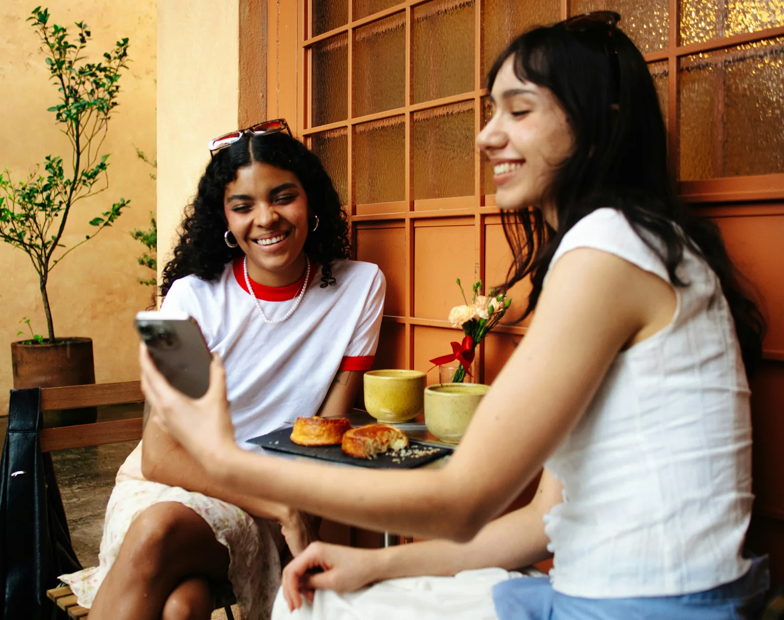 Two friends laughing at a café table, looking at a phone together